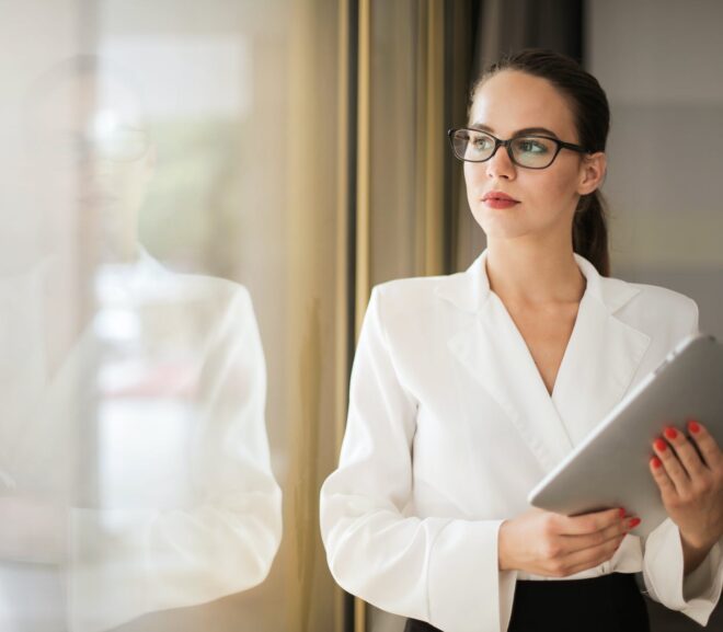 photo of woman in white top and glasses holding a tablet while looking outside a window